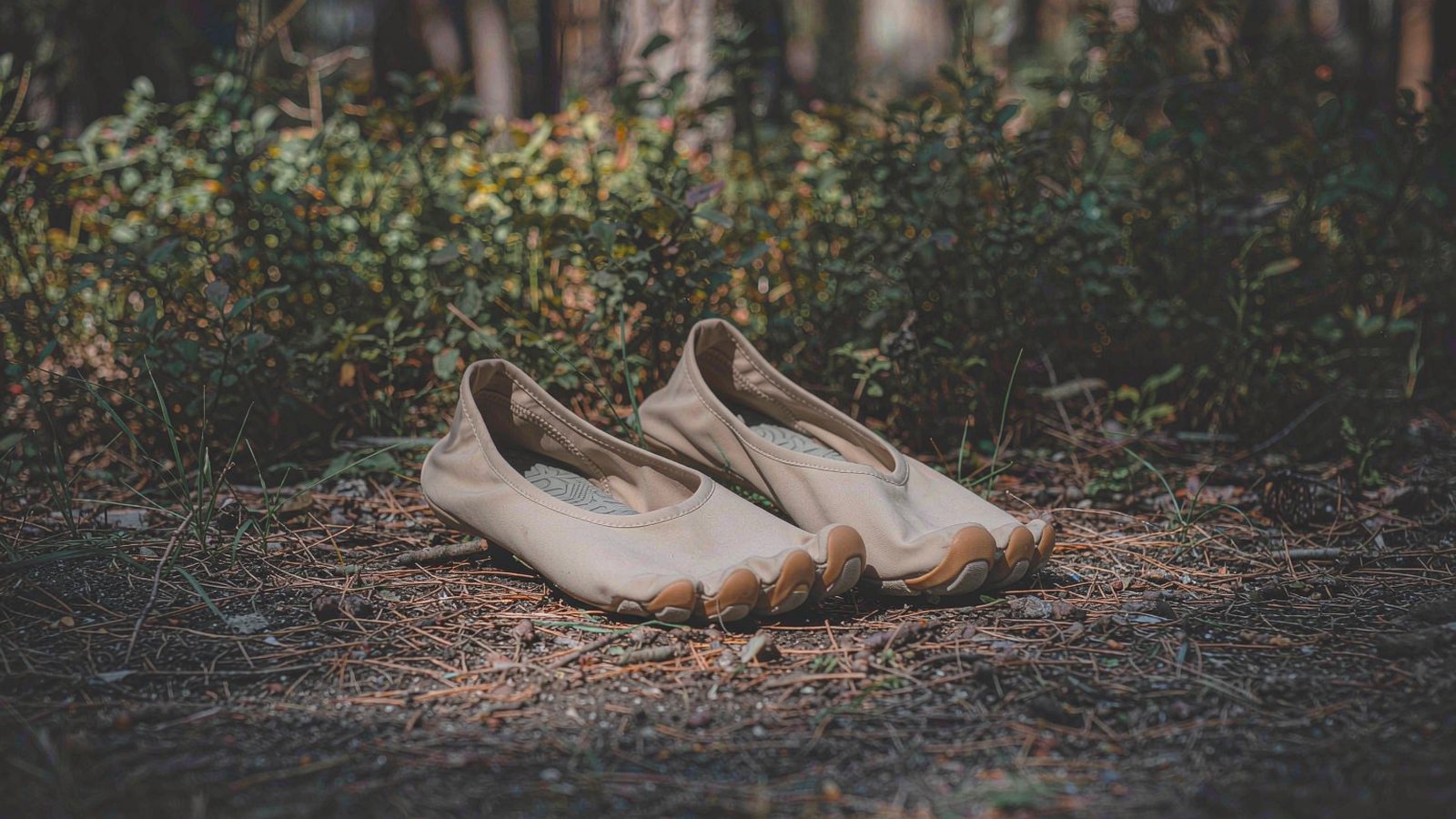 Thin, flexible barefoot shoes standing on a forest floor covered in pine needles under soft dappled light.
