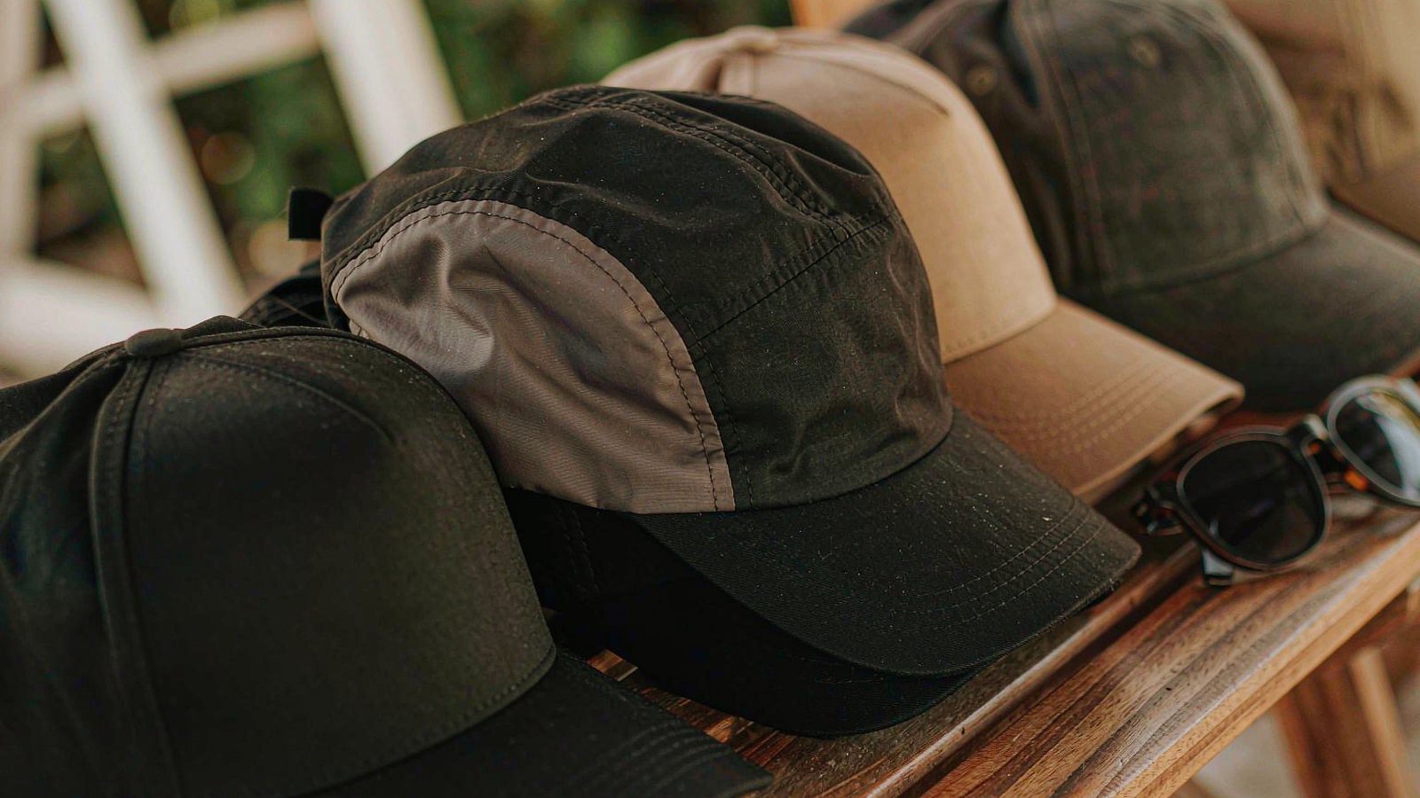 Several plain baseball caps stacked on a weathered wooden bench beside a pair of sunglasses, shot in warm daylight.