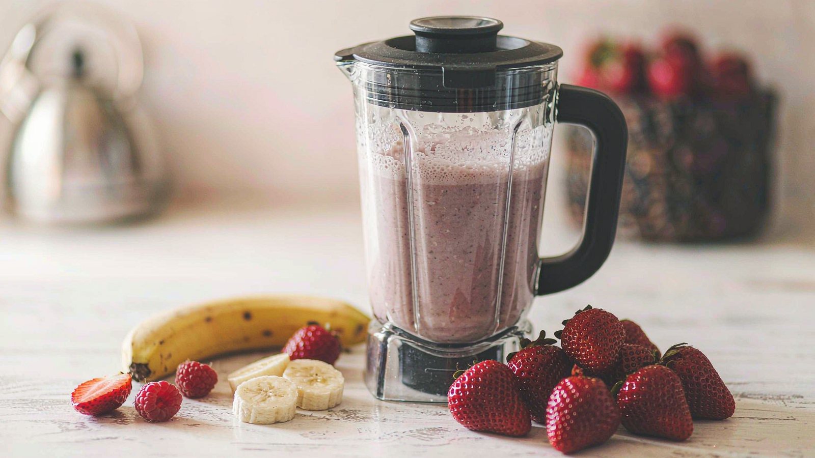 A blender filled with a berry smoothie on a bright kitchen counter with fresh fruit nearby in natural light