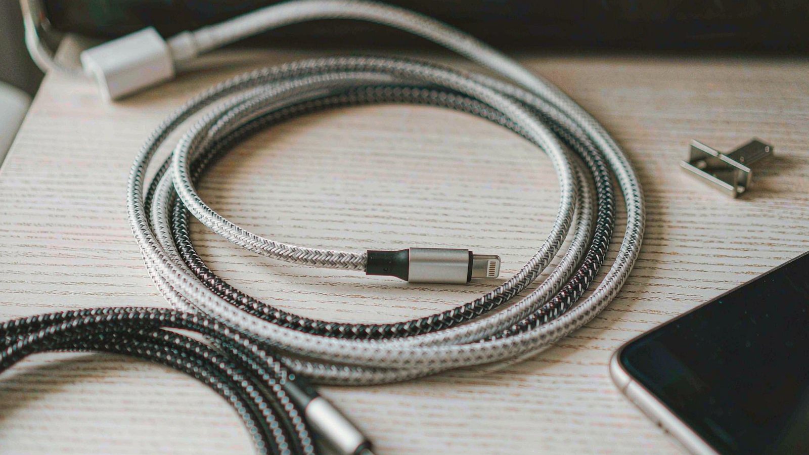 Several braided charging cables neatly coiled on a pale wooden surface beside a phone in soft daylight