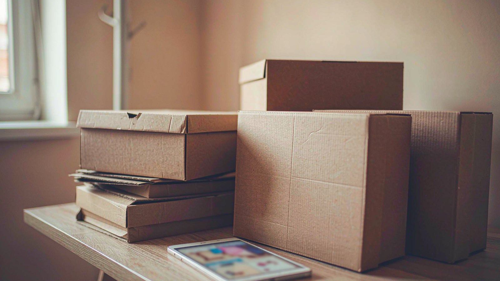 Stacked sneaker boxes on a wooden floor with a phone showing a blurred shopping page beside them in warm indoor light