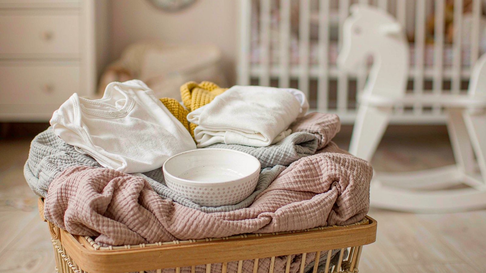 A gift basket of soft muslin blankets and small practical baby items arranged on a pale nursery floor in warm, gentle daylight