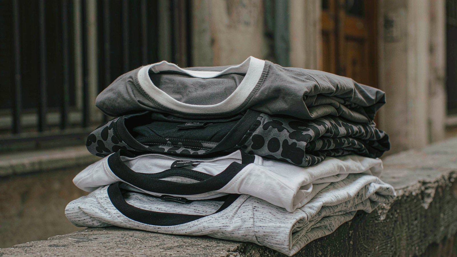 Folded graphic t-shirts stacked on a concrete ledge in a narrow alley under soft daylight