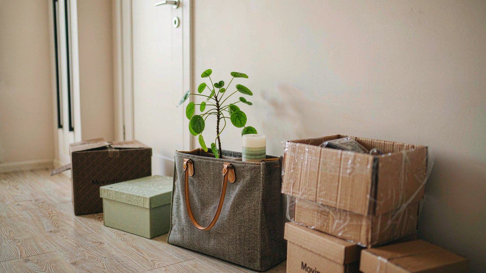 A housewarming gift bag with a small potted plant and candles resting on a wooden floor beside partially unpacked moving boxes in soft daylight