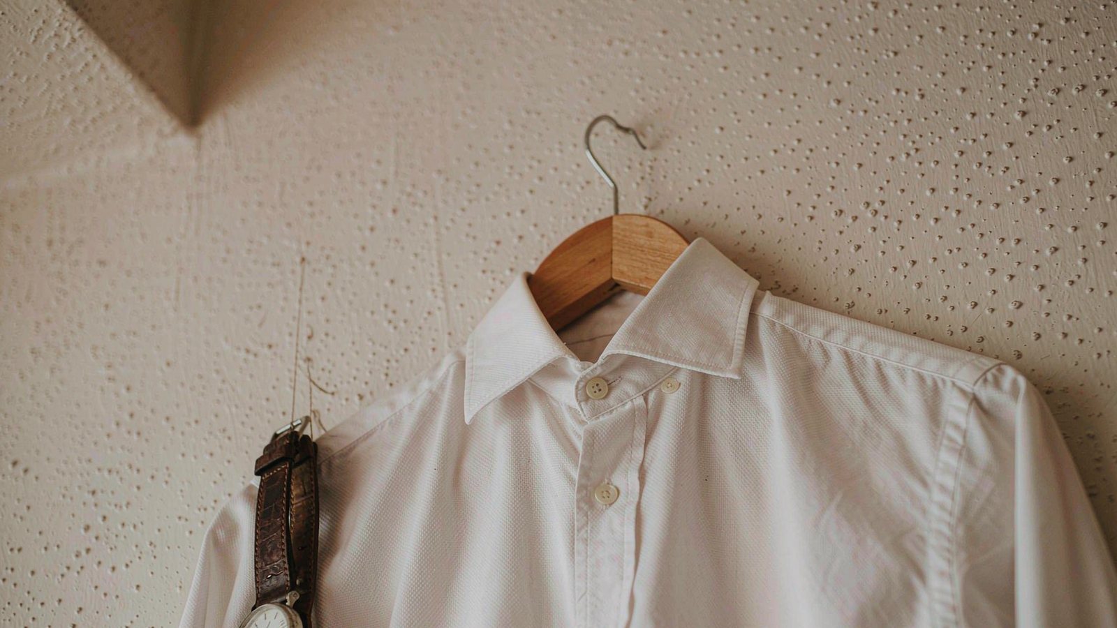 A crisp white Oxford shirt on a wooden hanger against a plaster wall with a leather watch resting below in soft side light