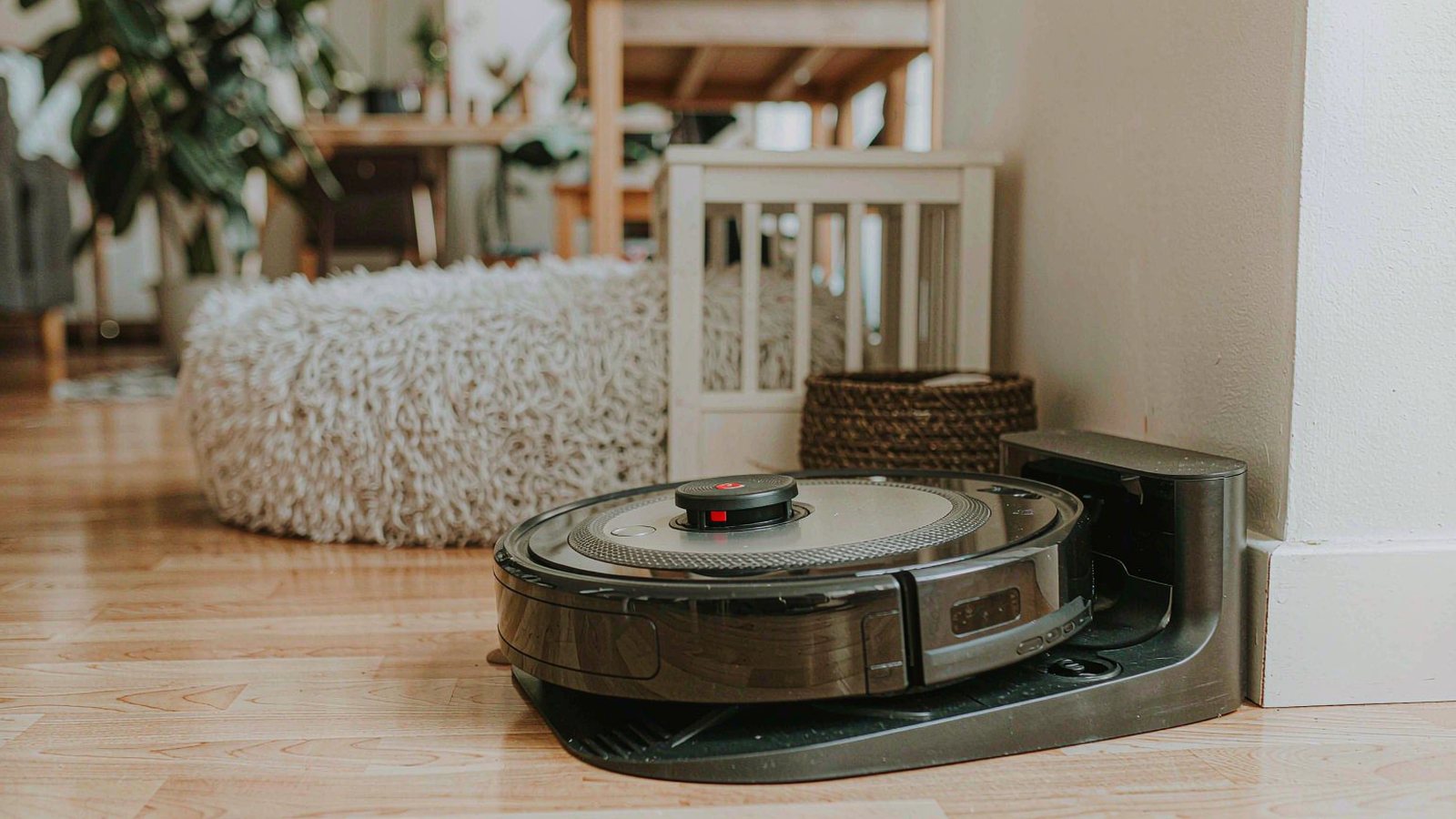 A round robot vacuum docked on light hardwood floor in a bright modern living room with a dog bed blurred in the background
