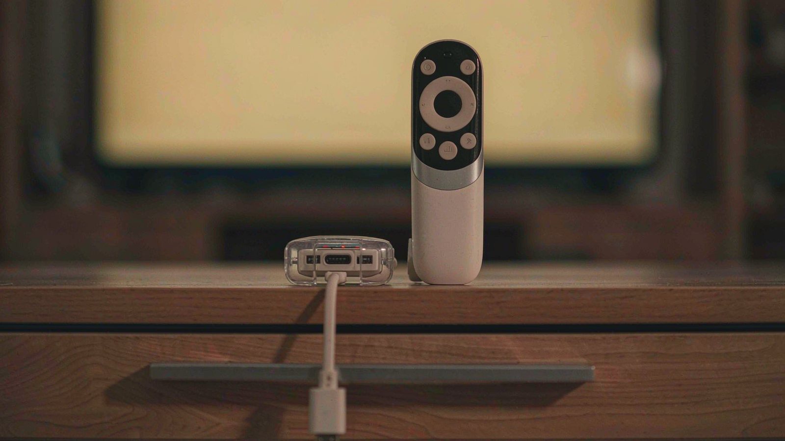 A small streaming stick and its remote resting on a wooden media console in front of a softly glowing television in dim evening light