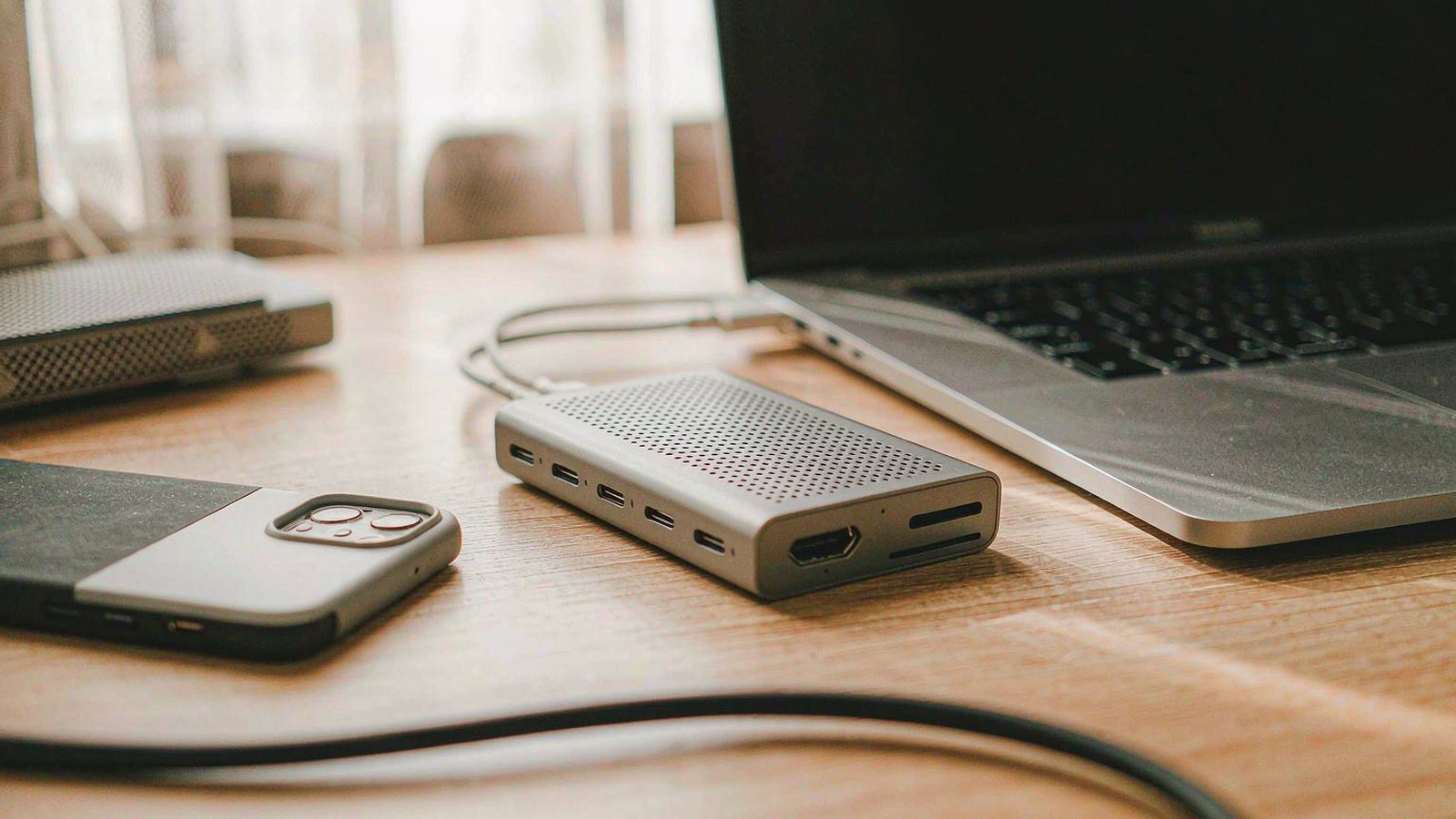 A compact aluminium USB-C hub plugged into a thin laptop on a tidy wooden desk with cables routed neatly behind, warm indoor light