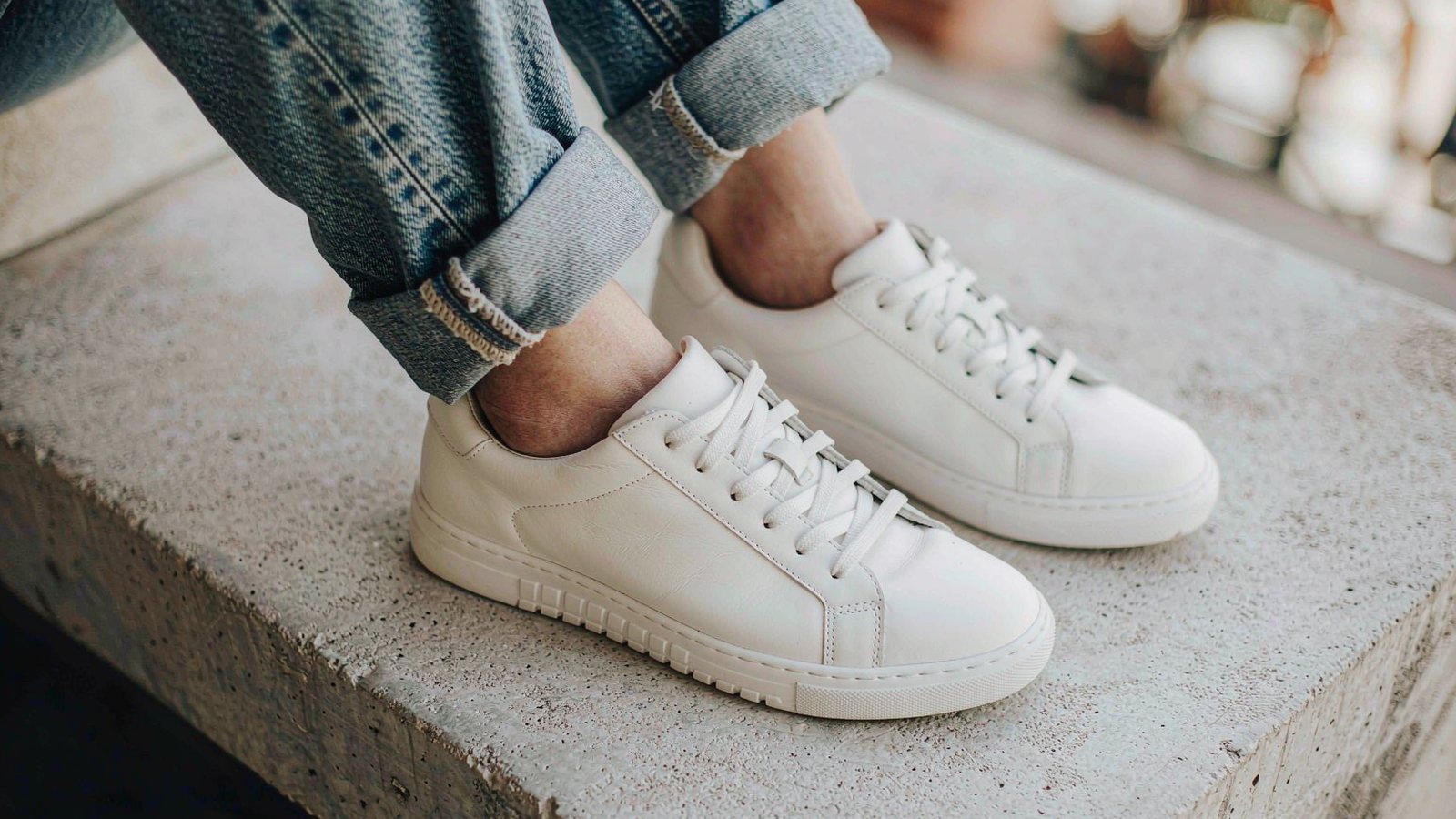 Clean white leather sneakers placed on a concrete step beside rolled-up jean hems, photographed in bright natural daylight.