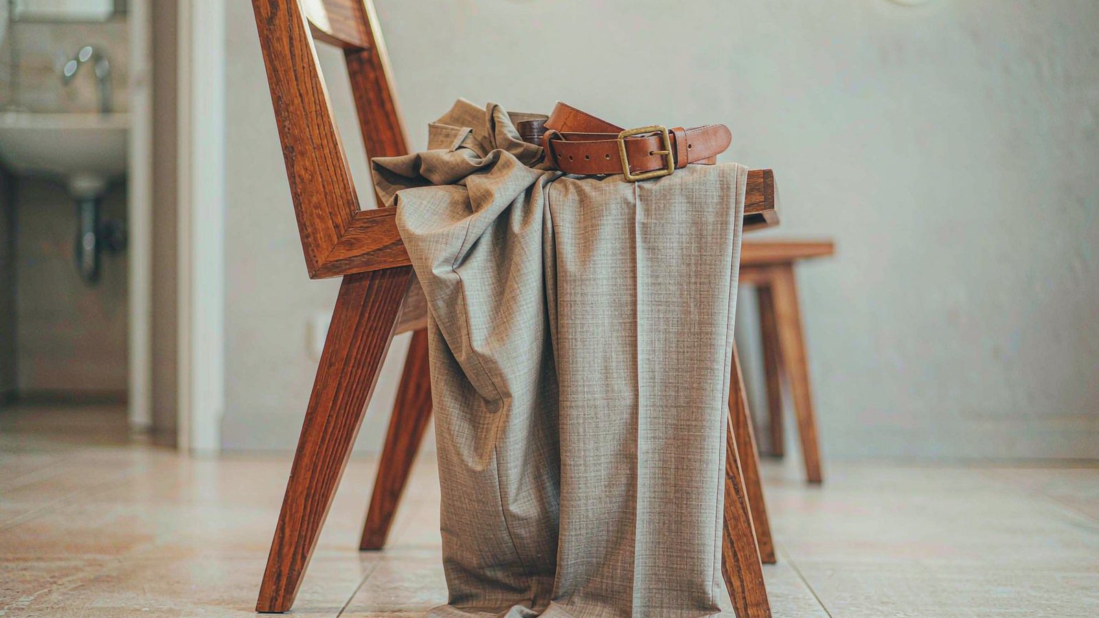 Tailored neutral trousers draped over a wooden chair with a leather belt in a bright minimal room